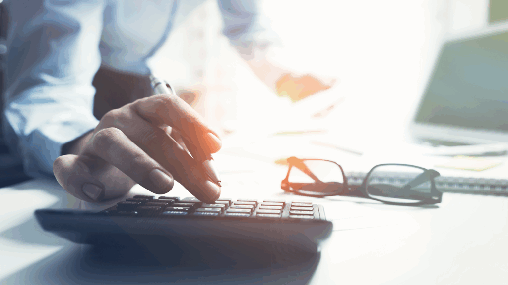 Person using a calculator on a desk with glasses and documents, possibly calculating temporary tax.